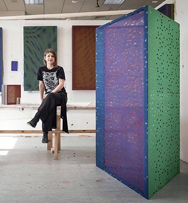 image of Gina Hunt, Director of the Ralph Arnold Gallery, seated on a stool in a studio setting with artwork in the foreground and background